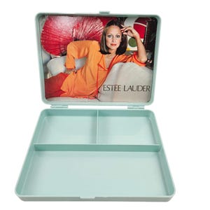 May include: A vintage, pale blue lunchbox, open to reveal a divided interior. The lid displays a retro advertisement with a woman in an orange robe, and the text "ESTEE LAUDER".
