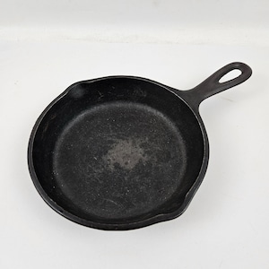 May include: A black cast iron skillet with a handle and a small pouring spout. The skillet is round and has a well-worn appearance, suggesting frequent use. The handle has a hole for hanging.