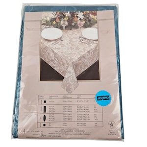 May include: A packaged tablecloth with a floral design, visible through the clear plastic. The tablecloth is beige with a detailed pattern, and the packaging includes size and care information. The package is teal.