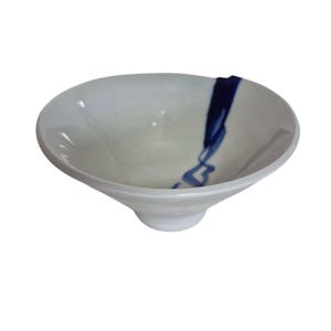 May include: White ceramic bowl with a flared rim and a small footed base. A dark blue, abstract design flows down the side of the bowl. The bowl has a smooth, glossy finish and is set against a white background.