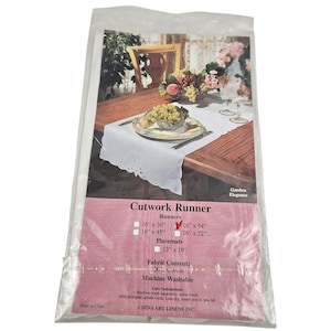 May include: A packaged white cutwork runner with a floral design, shown on a wooden table setting. The package also includes placemats. The text on the package reads "Cutwork Runner" and lists runner sizes.