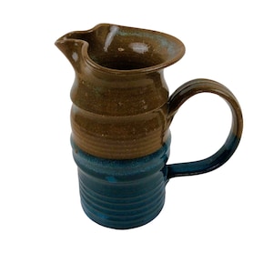 May include: A brown and blue ceramic pitcher with a wide mouth and a handle. The pitcher has a rustic, handmade look.