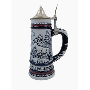 May include: A white ceramic beer stein with a blue and red trim. The stein features a detailed illustration of two moose in a forest setting. The lid is silver with a hinged handle. The text on the stein reads "Dall Sheep Alaskan Moose Fair"