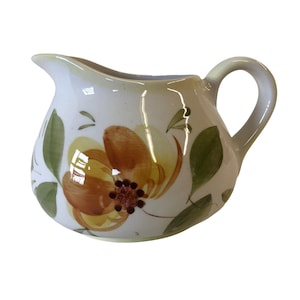 May include: A white ceramic creamer with a yellow and brown floral design. The creamer has a rounded body and a handle.