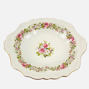 Puede incluir: Un cuenco de servir redondo de porcelana color crema con borde festoneado y ribete dorado. El cuenco presenta un patrón floral de rosas rosas y blancas con hojas verdes y pequeñas flores moradas dispuestas en un ramo central y un borde.