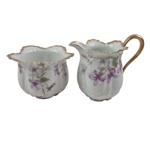 May include: A white porcelain creamer and sugar bowl set. The set features a floral design with purple flowers and green leaves. The rims and handle are trimmed with gold. The sugar bowl is 4 inches wide.