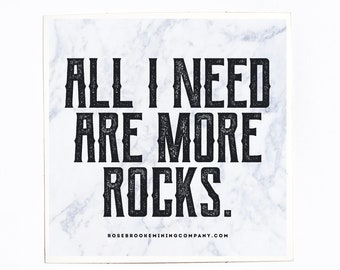 May include: Square sticker with a marble-effect background and the text "ALL I NEED ARE MORE ROCKS." The sticker also includes the website ROSEBROOKEMININGCOMPANY.COM. A wooden ruler is placed below the sticker.