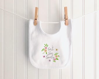 Könnte beinhalten: Ein weißes Baby-Lätzchen mit floralem Design und dem Text "isn't she lovely". Das Design umfasst rosa und gelbe Blumen, grüne Blätter und Schmetterlinge. Das Lätzchen hängt an einer Wäscheleine mit Holzklammern.