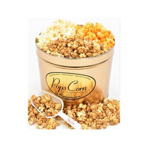 May include: A gold tin filled with three different flavors of popcorn: caramel, cheddar, and white cheddar. The tin has the words "Pops Corn" and "America's Finest Flavored Popcorn" printed on it. The tin is sitting on a white surface with a white scoop filled with caramel popcorn.