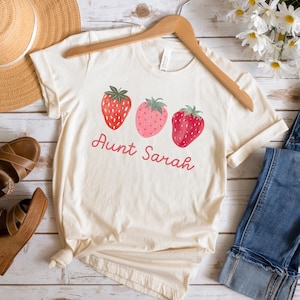 May include: Cream-colored t-shirt featuring three strawberry illustrations in shades of red and pink, with green leaves. The text "Aunt Sarah" is written in a cursive font below the strawberries. The shirt is displayed on a wooden hanger.