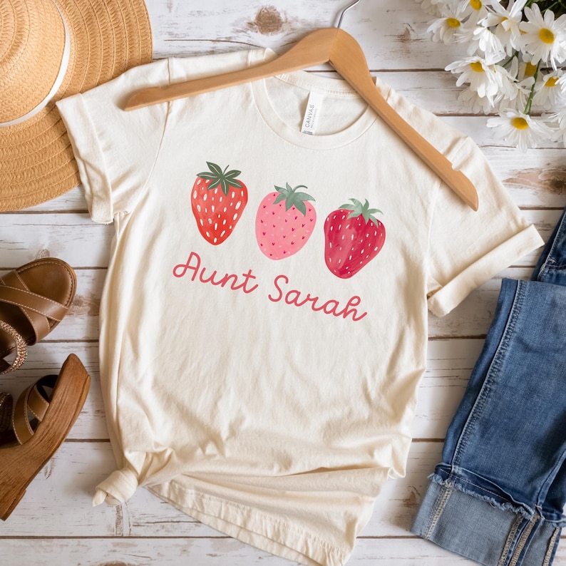 May include: Cream-colored t-shirt featuring three strawberry illustrations in shades of red and pink, with green leaves. The text "Aunt Sarah" is written in a cursive font below the strawberries. The shirt is displayed on a wooden hanger.
