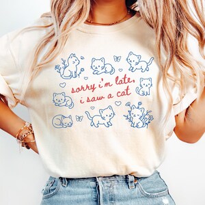 May include: Ivory-colored t-shirt featuring a playful design of blue cat illustrations, butterflies, and floral accents. The text "sorry i'm late, i saw a cat" is written in red. The shirt is made of a soft material.