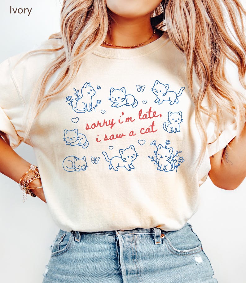 May include: Ivory-colored t-shirt featuring a playful design of blue cat illustrations, butterflies, and floral accents. The text "sorry i'm late, i saw a cat" is written in red. The shirt is made of a soft material.