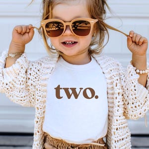 May include: A young child wearing a white t-shirt with the word "two" printed in brown. The child is wearing a white crocheted cardigan, brown shorts, and sunglasses. The child has a flower in their hair.