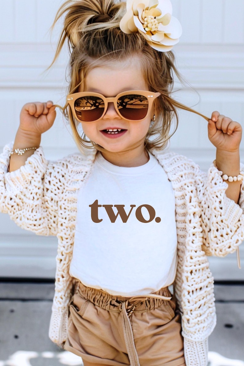 May include: A young child wearing a white t-shirt with the word "two" printed in brown. The child is wearing a white crocheted cardigan, brown shorts, and sunglasses. The child has a flower in their hair.
