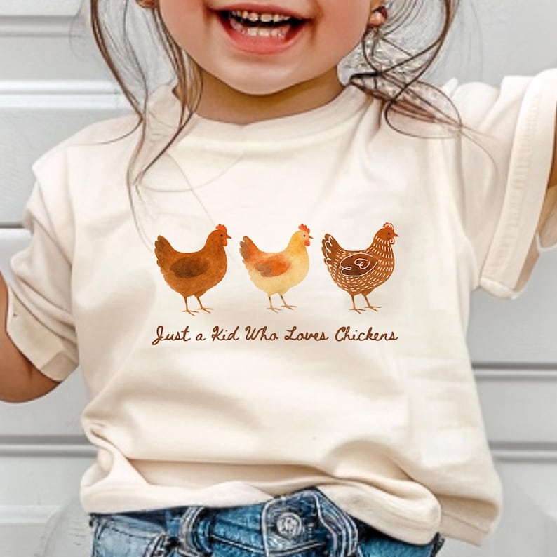May include: Cream-colored t-shirt featuring three watercolor chickens in shades of brown and yellow. The text "Just a Kid Who Loves Chickens" is printed below the chickens. The shirt is worn by a child.