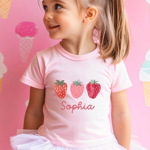 May include: A light pink t-shirt featuring three strawberry illustrations in varying shades of red and pink. The name "Sophia" is printed in a cursive font below the strawberries. The shirt is worn by a young child.