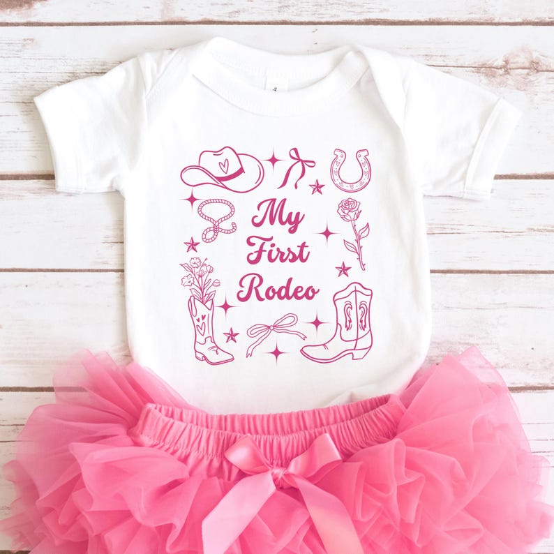May include: White baby onesie with pink cowboy-themed graphics, including a hat, boots, horseshoe, and the text "My First Rodeo".
