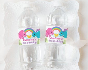 Può includere: Due bottiglie d'acqua in plastica trasparente con etichette personalizzate. Le etichette mostrano un dinosauro rosa e blu con un arcobaleno e il testo "Charlotte's 3rd Birthday".