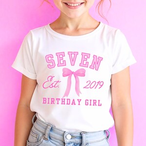 Peut inclure: T-shirt blanc avec un motif rose. Le motif comprend le mot "SEVEN" dans une police universitaire, un n&oelig;ud rose, le texte "Est. 2019" et les mots "BIRTHDAY GIRL". Le t-shirt est port&eacute; par une personne.