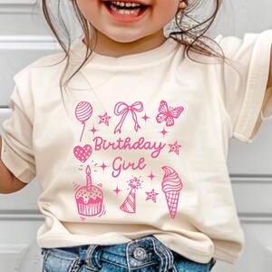 May include: A light beige t-shirt with a pink graphic design that says "Birthday Girl" and features a cupcake, ice cream cone, party hat, balloon, bow, butterfly, and stars.