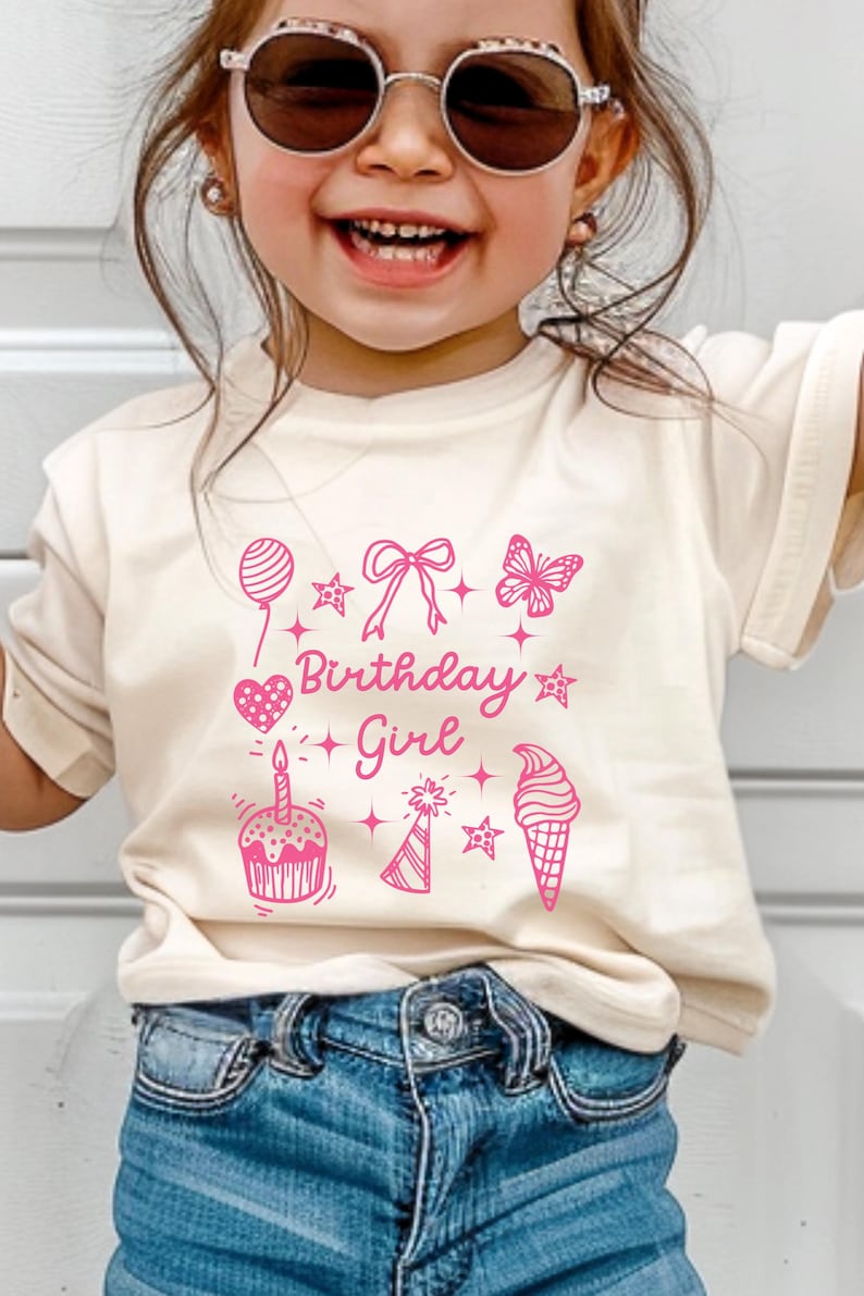 May include: A light beige t-shirt with a pink graphic design that says "Birthday Girl" and features a cupcake, ice cream cone, party hat, balloon, bow, butterfly, and stars.