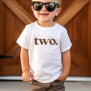 May include: A white t-shirt with brown text that says "two." A young child is wearing the shirt and has on black sunglasses.