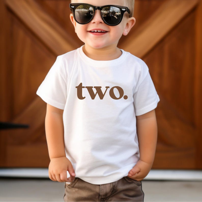 May include: A white t-shirt with brown text that says "two." A young child is wearing the shirt and has on black sunglasses.