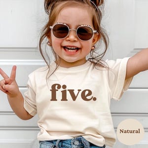 May include: A young child wearing a cream-colored t-shirt with the word "five" in brown lettering. The child is also wearing sunglasses and smiling. The shirt is labeled "Natural" in a small circle.
