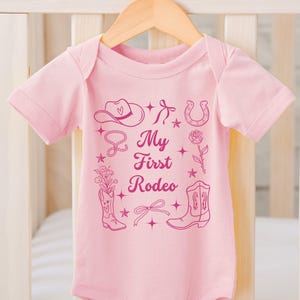 May include: Pink baby onesie with a pink graphic of a cowboy hat, boots, horseshoe, lasso, and a rose. The text "My First Rodeo" is printed on the onesie.