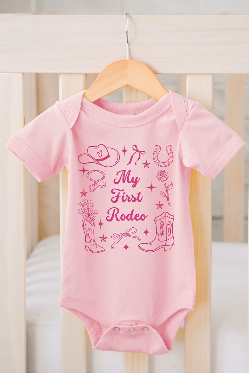 May include: Pink baby onesie with a pink graphic of a cowboy hat, boots, horseshoe, lasso, and a rose. The text "My First Rodeo" is printed on the onesie.