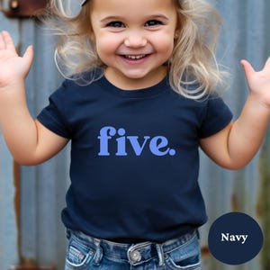 May include: Navy blue t-shirt with the word "five" in light blue. The shirt is worn by a child. The shirt is paired with blue jeans. The word "Navy" is in a circle in the bottom right corner.