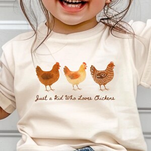 May include: Cream-colored t-shirt featuring three watercolor chickens in shades of brown and orange. The text "Just a Kid Who Loves Chickens" is printed below the chickens. The shirt is worn by a child.