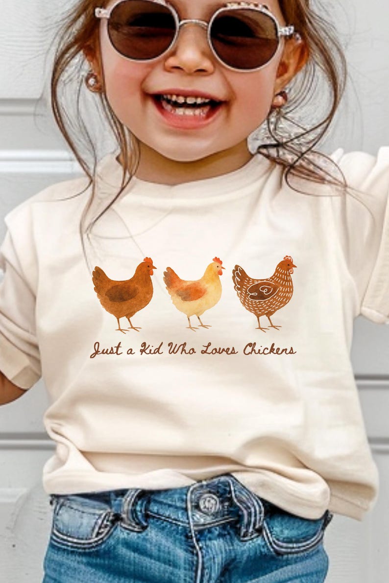 May include: Cream-colored t-shirt featuring three watercolor chickens in shades of brown and orange. The text "Just a Kid Who Loves Chickens" is printed below the chickens. The shirt is worn by a child.