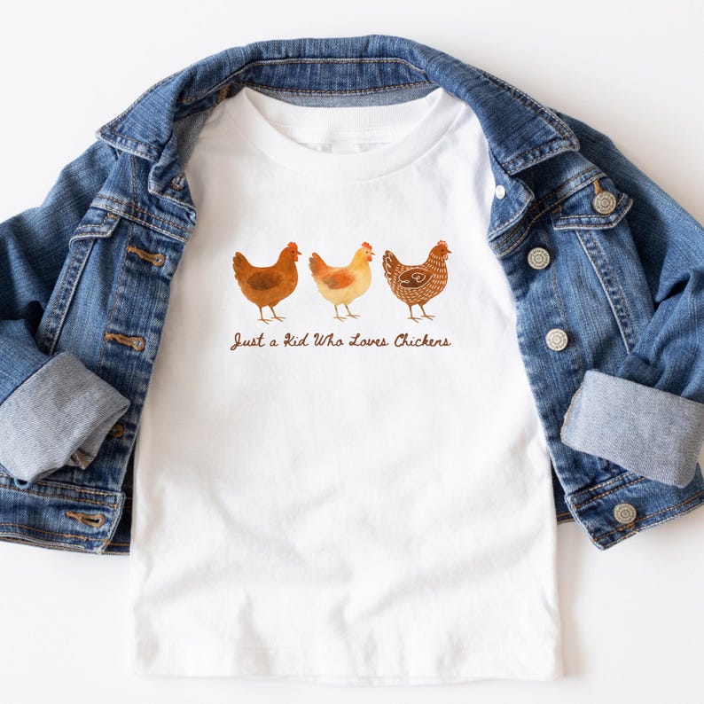 May include: White t-shirt featuring three watercolor chickens in shades of brown and yellow. The text "Just a Kid Who Loves Chickens" is printed below the chickens. The shirt is displayed on a denim jacket.
