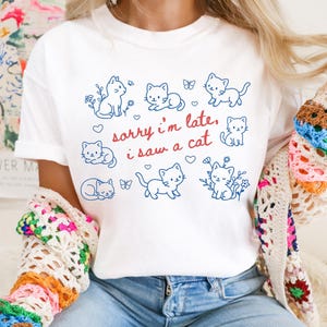 May include: White t-shirt featuring a blue outline design of several cats, flowers, and butterflies. The text "sorry i'm late, i saw a cat" is written in red script. The shirt is a casual, everyday wear item.