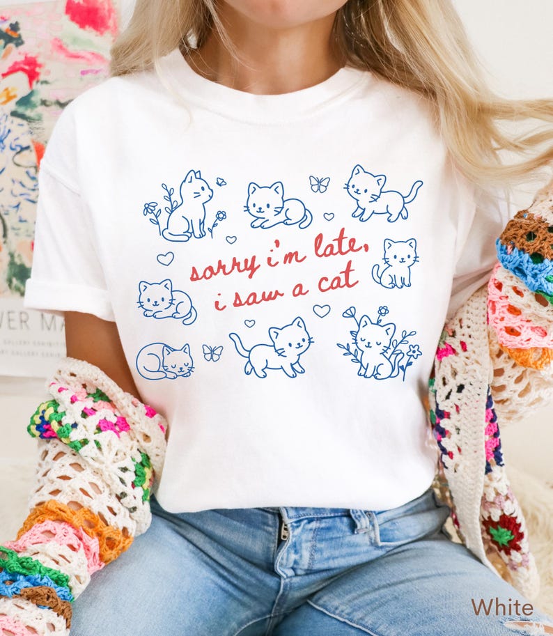 May include: White t-shirt featuring a blue outline design of several cats, flowers, and butterflies. The text "sorry i'm late, i saw a cat" is written in red script. The shirt is a casual, everyday wear item.