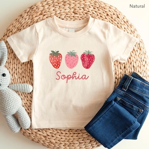 May include: A cream-colored t-shirt featuring three strawberry illustrations in red and pink, with green leaves. The name "Sophia" is written in a cursive font below the strawberries. Dark blue jeans are folded next to the shirt.