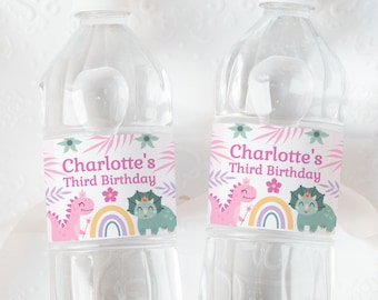 Può includere: Due bottiglie d'acqua in plastica trasparente con etichette a tema dinosauri rosa e verdi. Le etichette recitano "Charlotte's Third Birthday" con un dinosauro rosa, un dinosauro verde e un arcobaleno.
