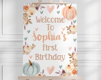 May include: Welcome to Sophia's First Birthday sign with a white background, floral designs, and two pumpkins. The pumpkins are one peach and one light blue. The text is in a copper color.