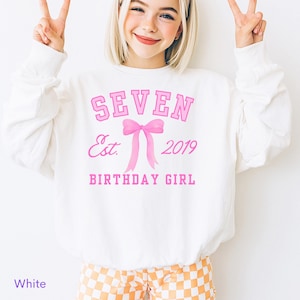 May include: White sweatshirt with the text "SEVEN Est. 2019 BIRTHDAY GIRL" in pink. A pink bow graphic is centered below the word "SEVEN". The sweatshirt is a crew neck style.