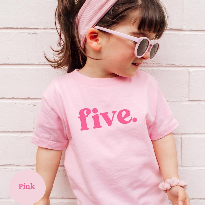 May include: A pink t-shirt with the word "five" in a darker pink font. The child is wearing a matching pink headband and sunglasses. A pink scrunchie is on the child's wrist. The background is a white brick wall.