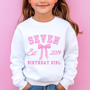Peut inclure: Sweat-shirt blanc avec texte rose : "SEVEN Est. 2019 BIRTHDAY GIRL" et un n&oelig;ud rose. Le sweat-shirt est de style col rond. Le fond est rose uni.