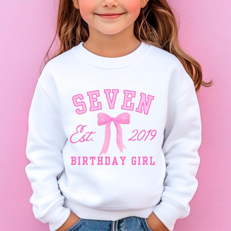 Peut inclure: Sweat-shirt blanc avec texte rose : "SEVEN Est. 2019 BIRTHDAY GIRL" et un n&oelig;ud rose. Le sweat-shirt est de style col rond. Le fond est rose uni.