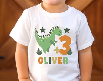 May include: White t-shirt with a green dinosaur illustration, the number 3 in orange, and the name "OLIVER" in colourful letters. The design includes green stars and foliage. A fun birthday shirt for a child.