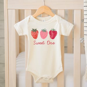 May include: Cream-colored baby bodysuit with three strawberry illustrations and the text "Sweet One" in a cursive font. The strawberries are red and pink with green leaves. The bodysuit is hanging on a wooden hanger.