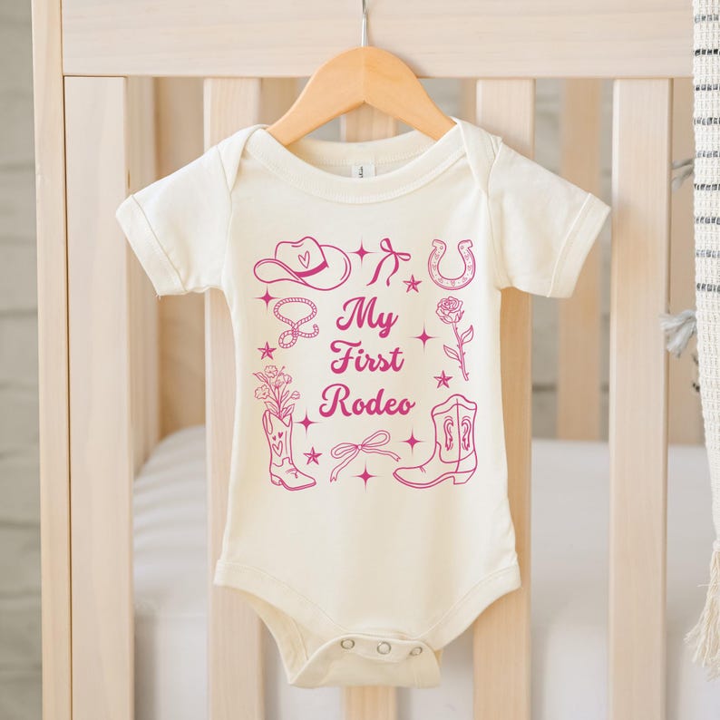May include: A white baby onesie with a pink design of a cowboy hat, boots, horseshoe, and a lasso. The text "My First Rodeo" is printed in pink on the onesie.