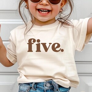 May include: A young child wearing a cream-colored t-shirt with the word "five" in brown lettering. The child is also wearing blue denim jeans and round sunglasses. The child is smiling.
