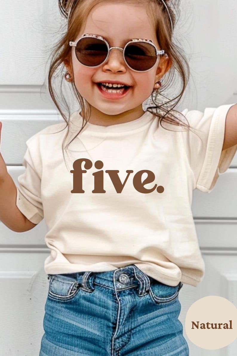 May include: A young child wearing a cream-colored t-shirt with the word "five" in brown lettering. The child is also wearing blue denim jeans and round sunglasses. The child is smiling.