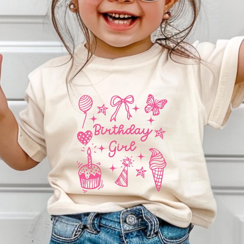 May include: A white t-shirt with a pink graphic design that says "Birthday Girl" and features a cupcake, ice cream cone, butterfly, balloon, and other birthday-themed illustrations.